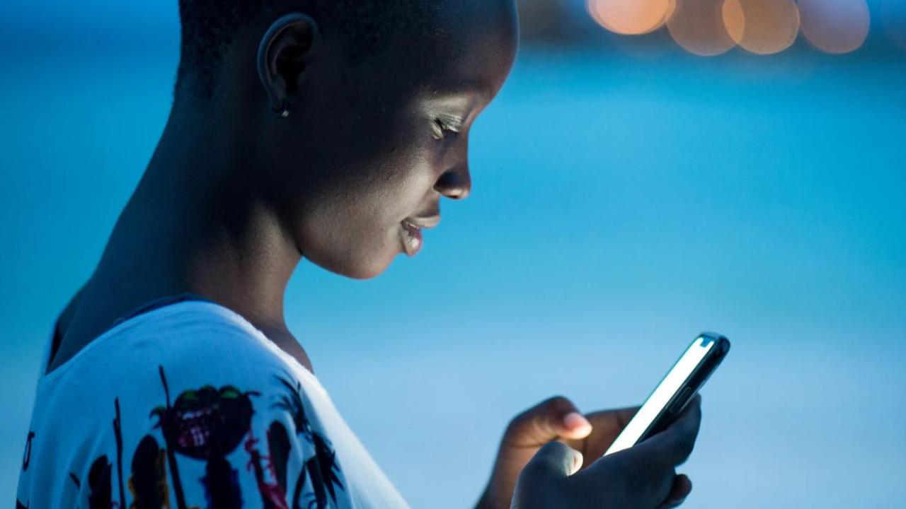 Close-up of a young woman using a smartphone at dusk, her face illuminated by the screen’s glow. She appears focused, with a soft blue background and blurred city lights in the distance.