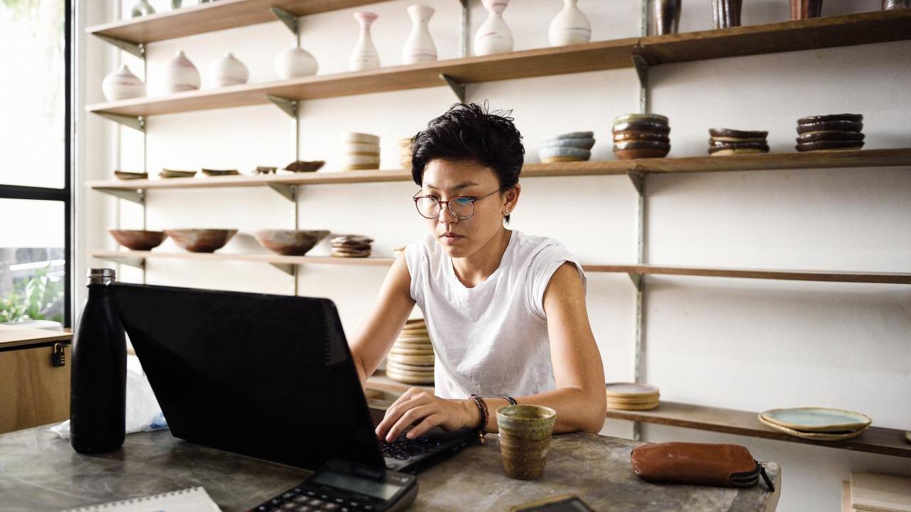 A person working on a laptop, shelves of ceramics behind them.