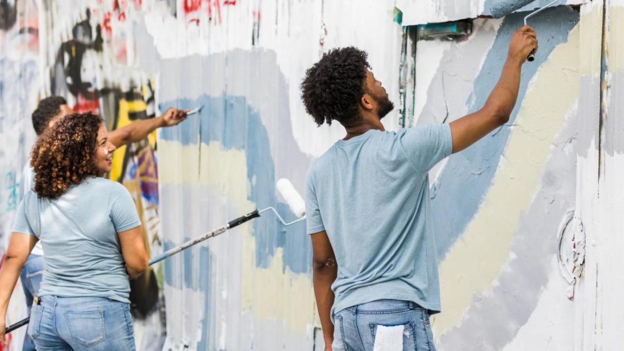 Group of young adults painting over a graffiti-covered wall with rollers and brushes, working together as part of a community mural or cleanup project. They wear casual clothes and appear engaged and cheerful.