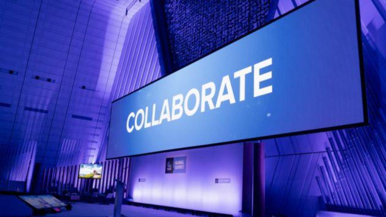  A large digital screen in a modern conference hall displays the word "COLLABORATE" in bold white letters against a blue background. The hall is illuminated with blue and purple lighting, creating a professional and dynamic atmosphere.