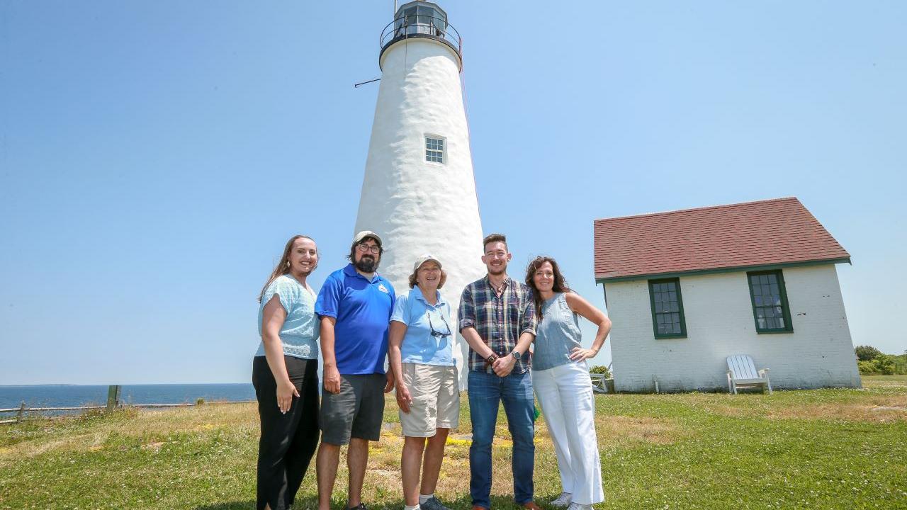 A group of five people pose in front of a light house on an island.