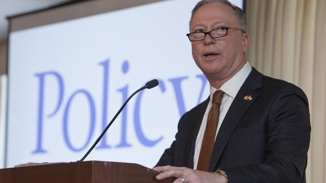 Greg Ebel at a podium with microphone. A screen behind him with "Policy" on it.