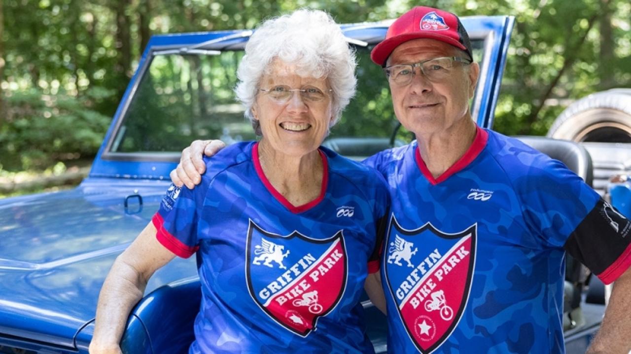 Gene and Donna Griffin, standing in front of a jeep, both wearing matching blue Griffin Bike Trail bike shirts
