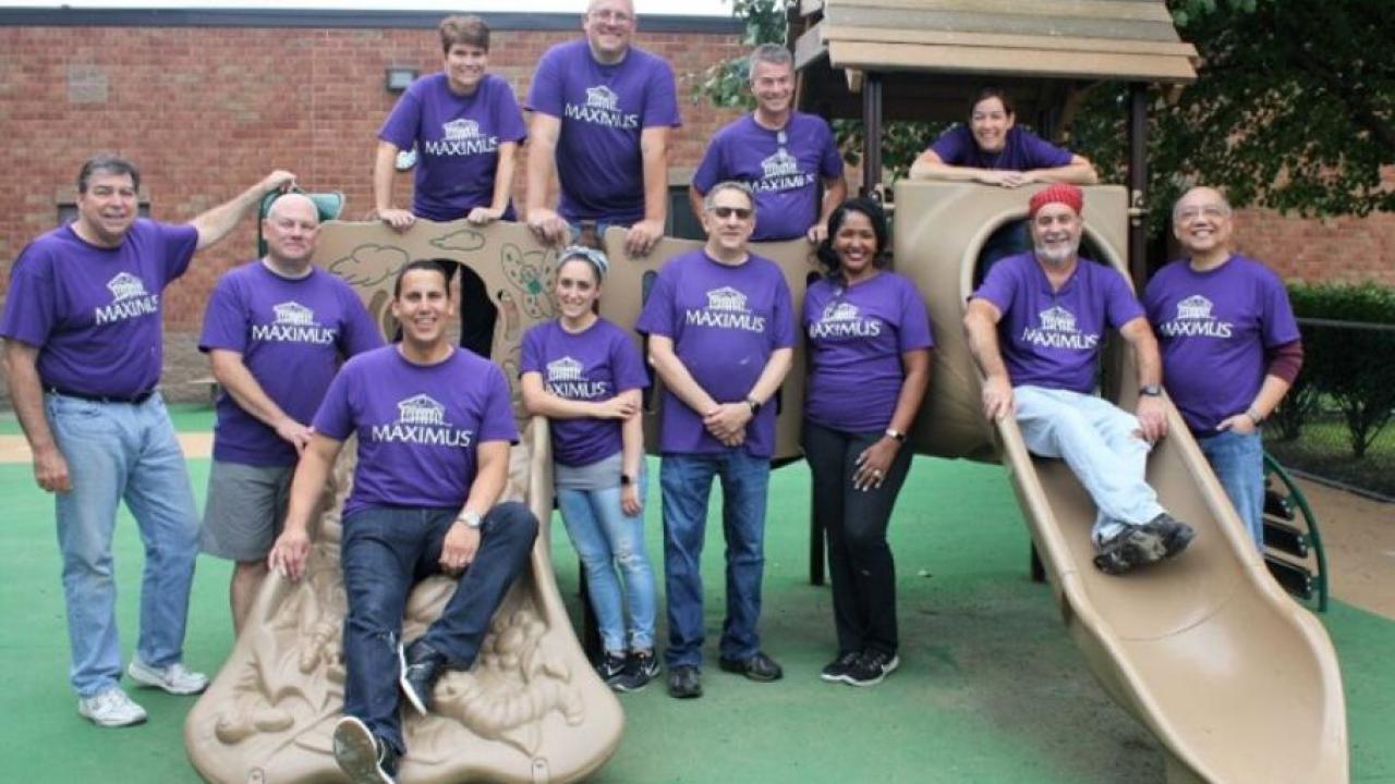 A group of people stood around a children's climbing frame, wearing purple MAXIMUS t-shirts
