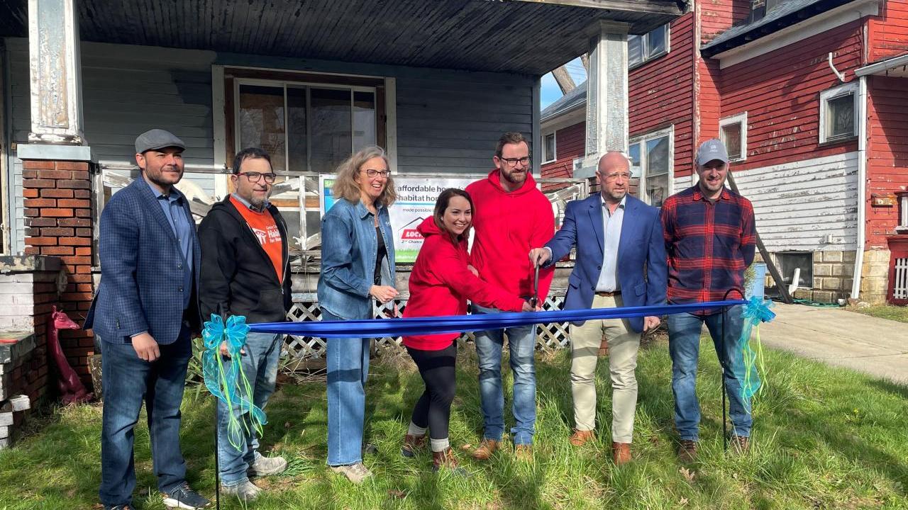 A group posed cutting a blue ribbon in front of a house.