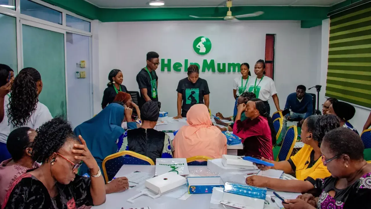 A group of people, some seated, some standing, in a conference room. A speaker at the front. "Help Mum" sign behind them.