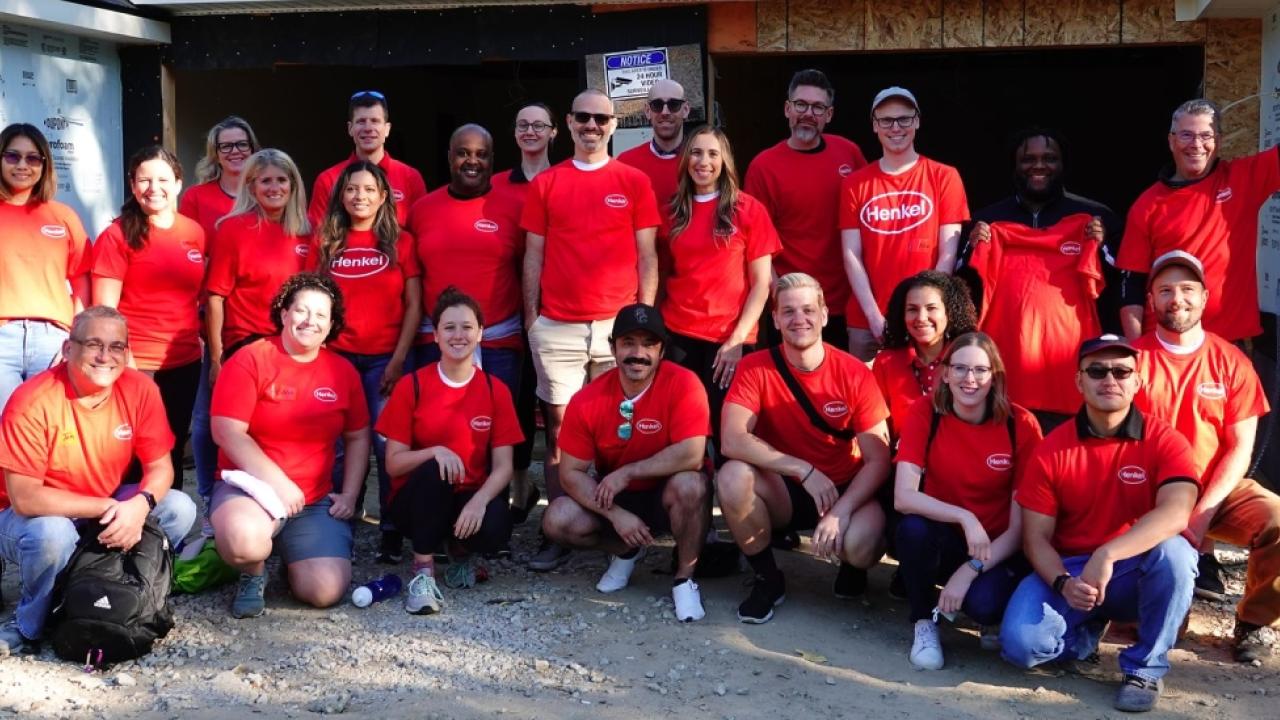 group of people outside a home being built, all are wearing matching orange shirts with Henkel logo
