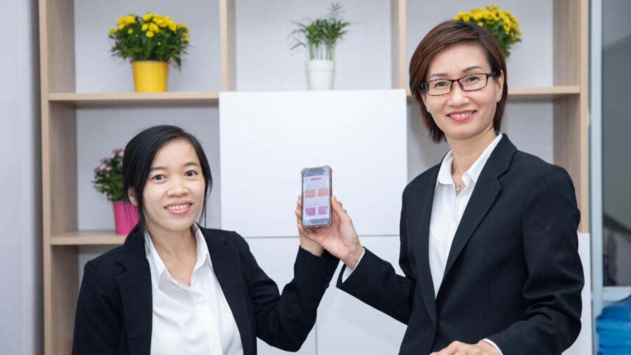 Two people holding a phone up. Shelves behind them each with flowers in them.
