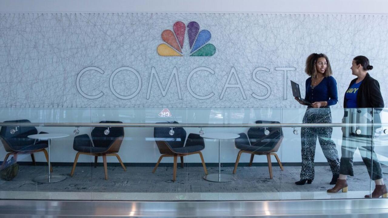 Two people walking a hallway in front of a large Comcast sign