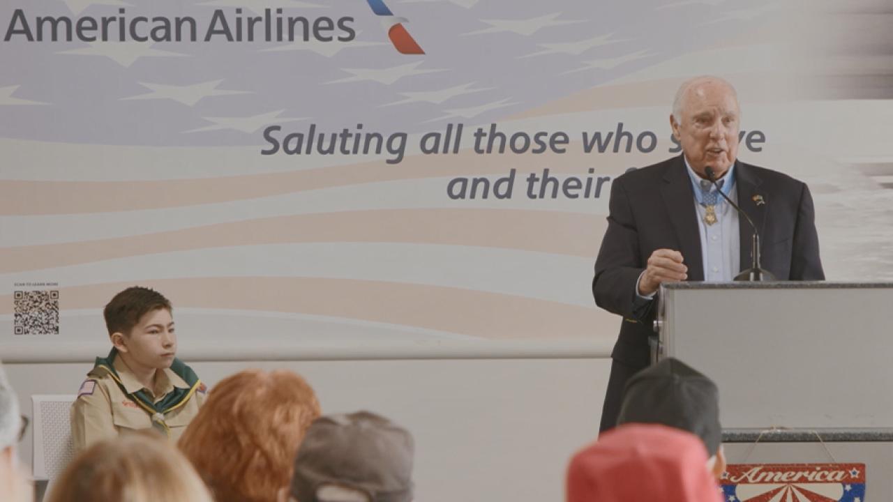 person stands at a podium in front of a crowd. American Airlines logo behind them