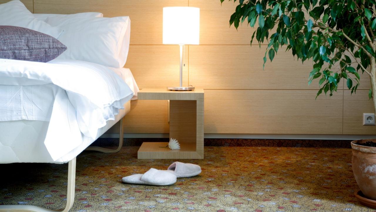 Horizontal view of a hotel room with a bed, side table, lamp, and slippers