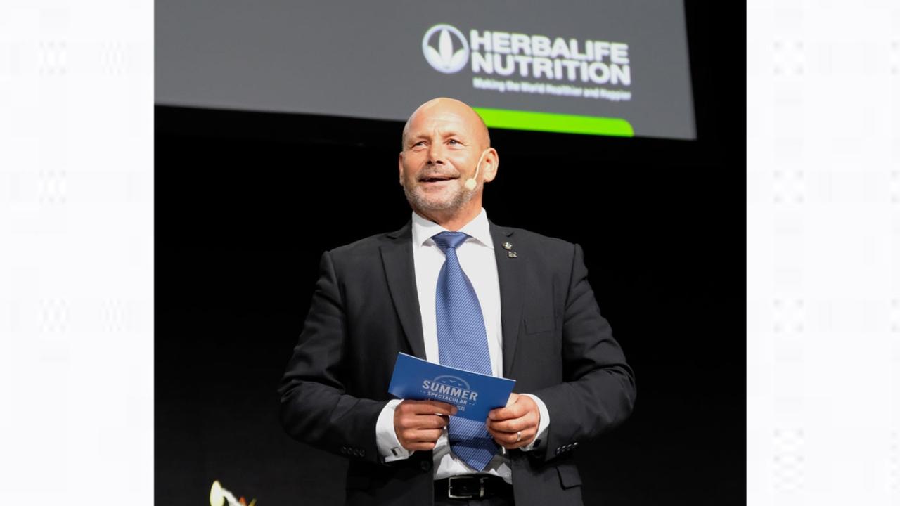 A person standing on a stage, holding a blue card. "herbalife nutrition" logo behind them.