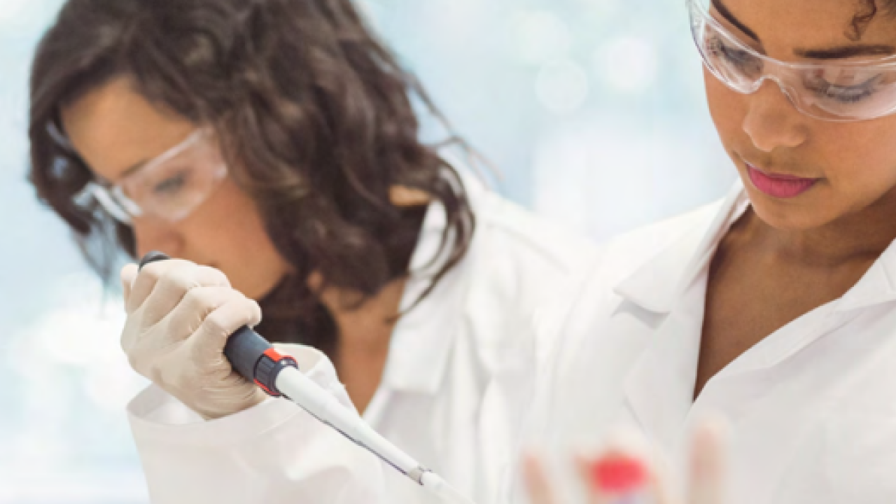 Three people in protective lab gear. One holds a pipette.