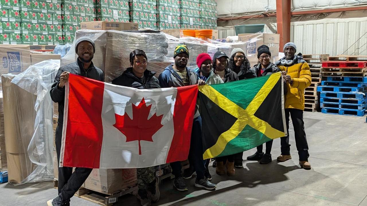 DP World employees and local volunteers stand inside a warehouse holding Canadian and Jamaican flags in front of palletized relief supplies bound for hurricane-affected communities in Jamaica.