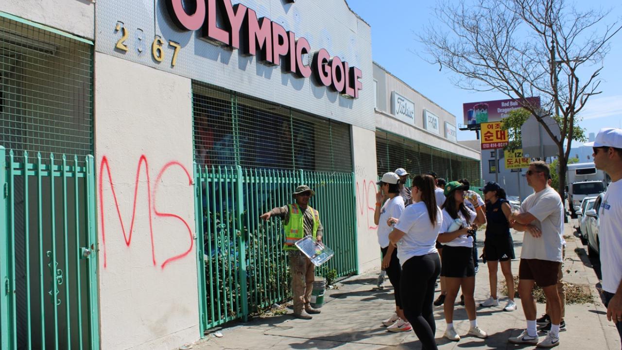 Community volunteers gather on a sidewalk as a KYCC cleanup leader provides instructions in front of a building marked with graffiti.