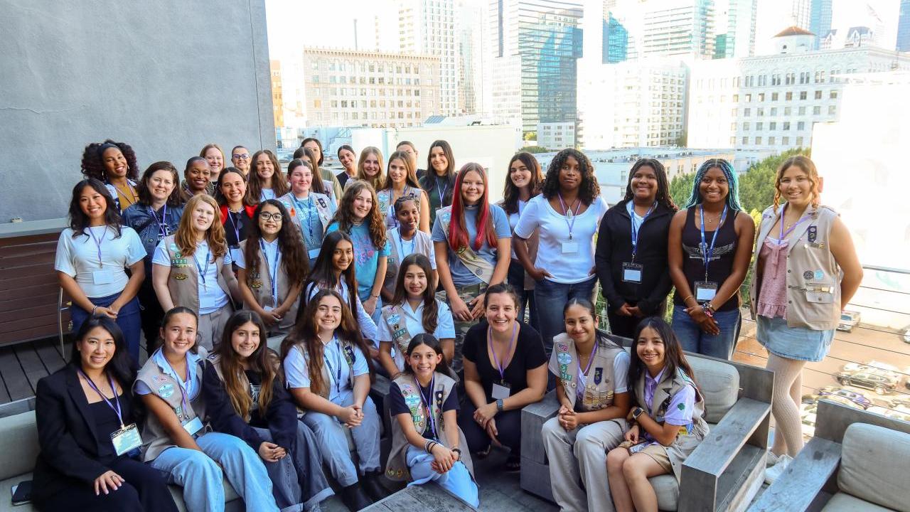A large group of participants gathers on an outdoor terrace with city buildings in the background, posing together during a group activity.