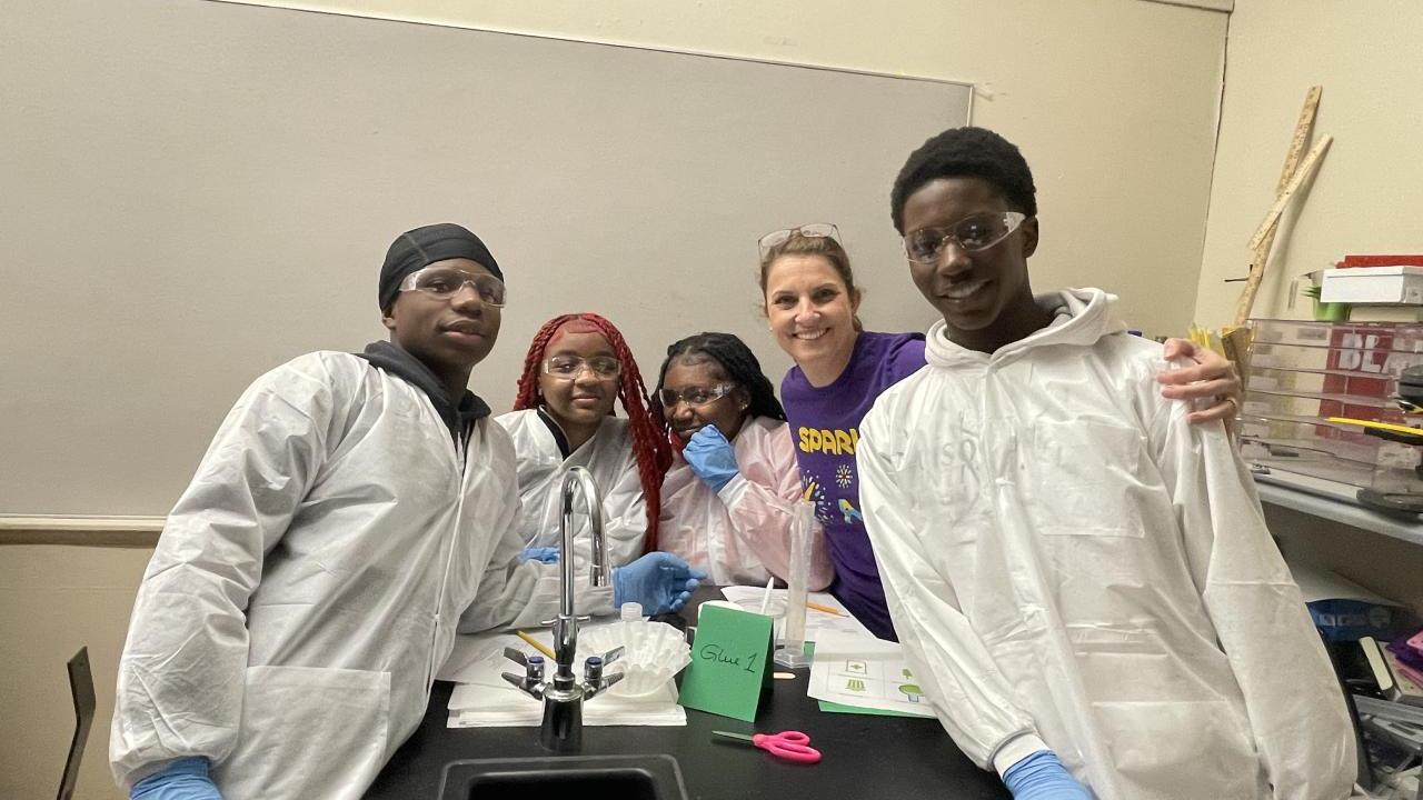 A woman in a purple Curiosity Labs™ MilliporeSigma t-shirt stands with students in lab equipment as they conduct experiments. 