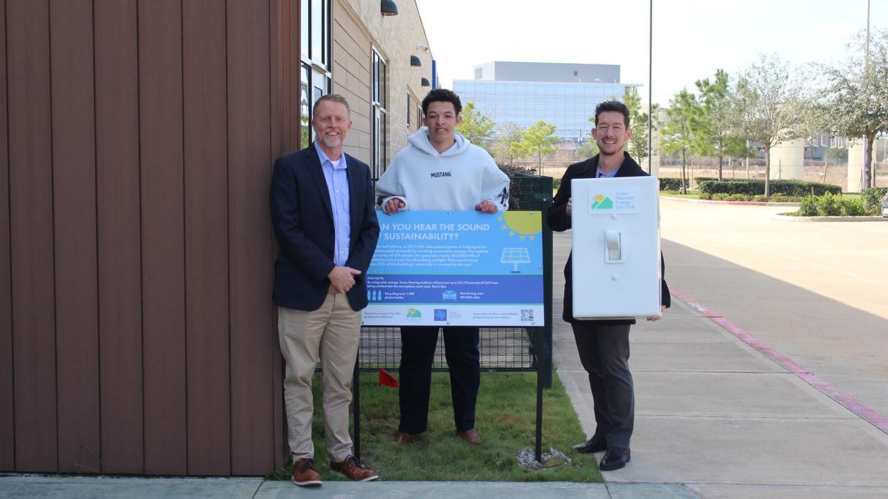 Three men standing around blue sign smiling, with one of them holding an oversized prop light switch