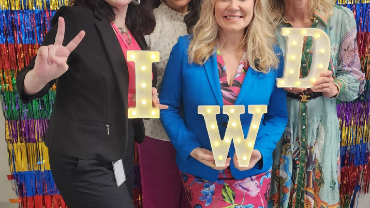Four people posed in front of a wall of colorful fringe. Three holding "IWD" letters.