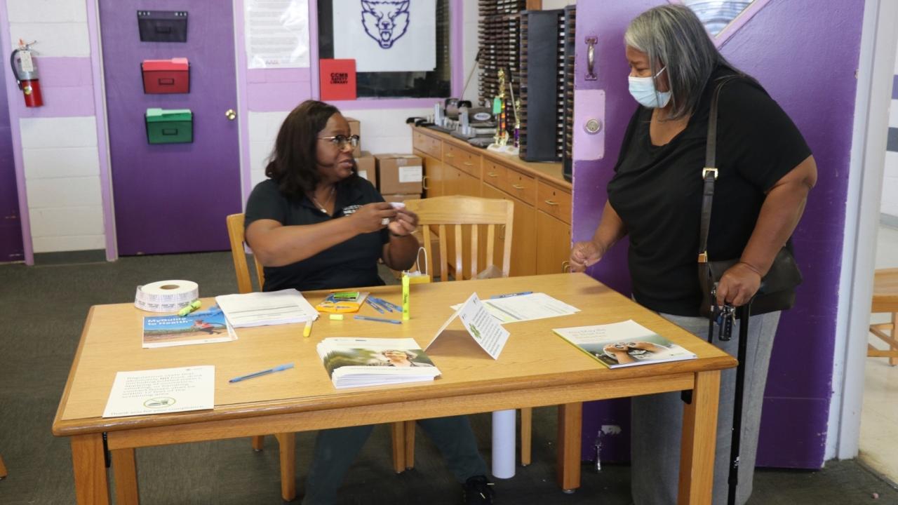 Stacey Ingram sitting at a table with stacks of literature. A person standing beside her wearing a protective mask.