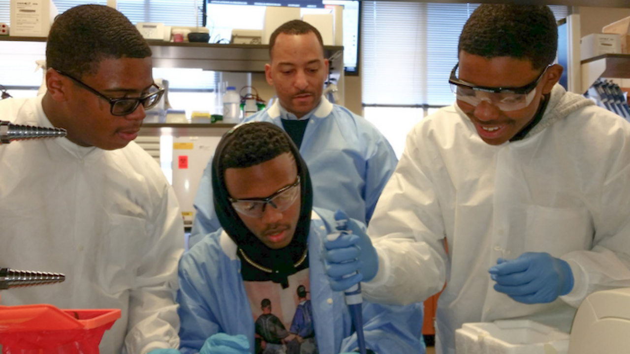 Dr. Arik King in a lab working with three young men of color. All are wearing lab coats and protective eye ware. 