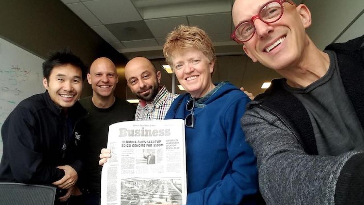 Shannon Whitmore with three men, holding a newspaper with a headline "Illumina Buys Startup"