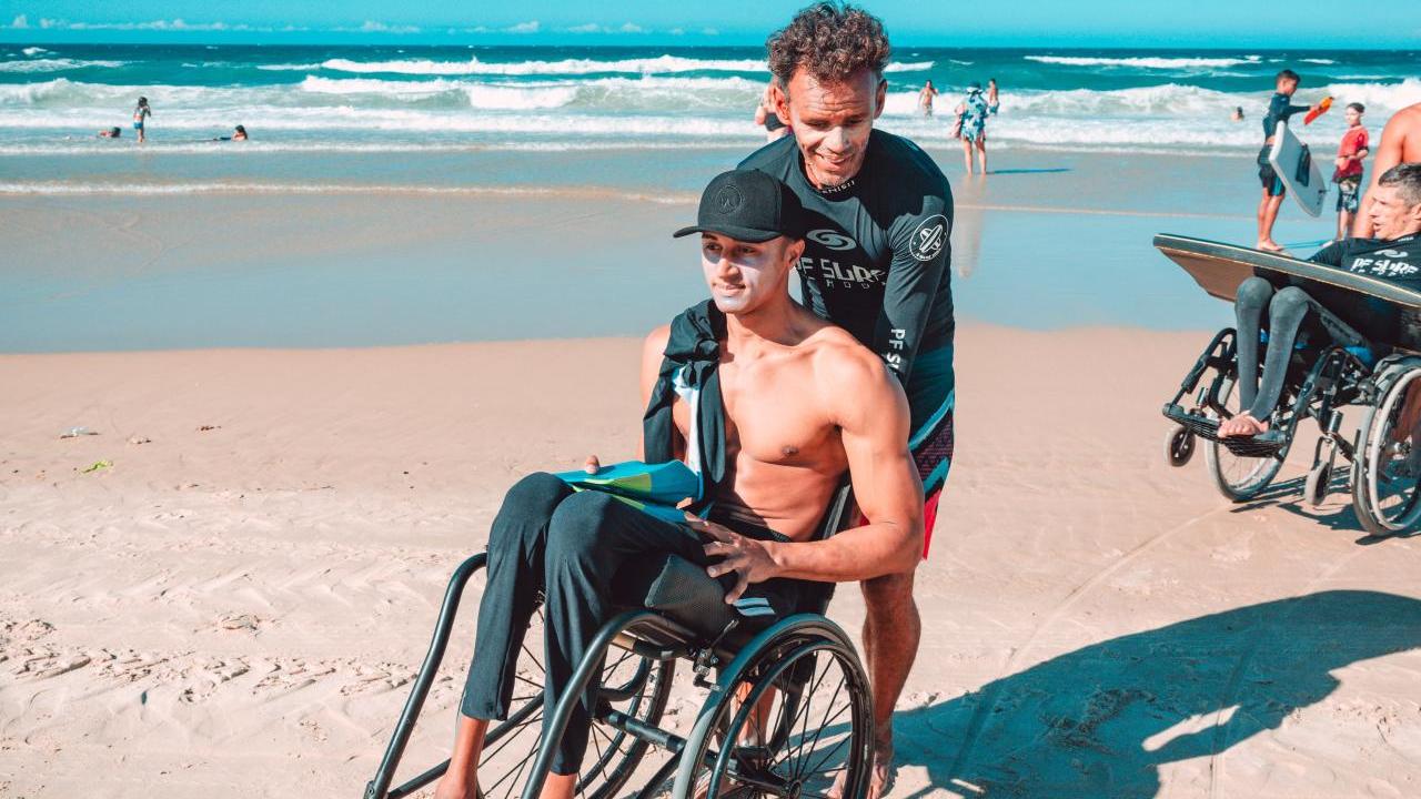 A person in a wet suit helping another in a wheelchair on a beach.