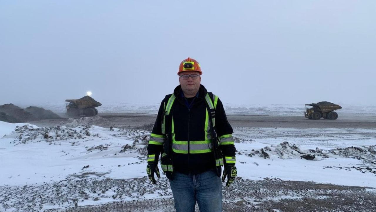 Jason Mercer in protective and high-vis jacket and hard hat outside in a snow covered quarry, dense fog behind them and two large mining dump trucks.