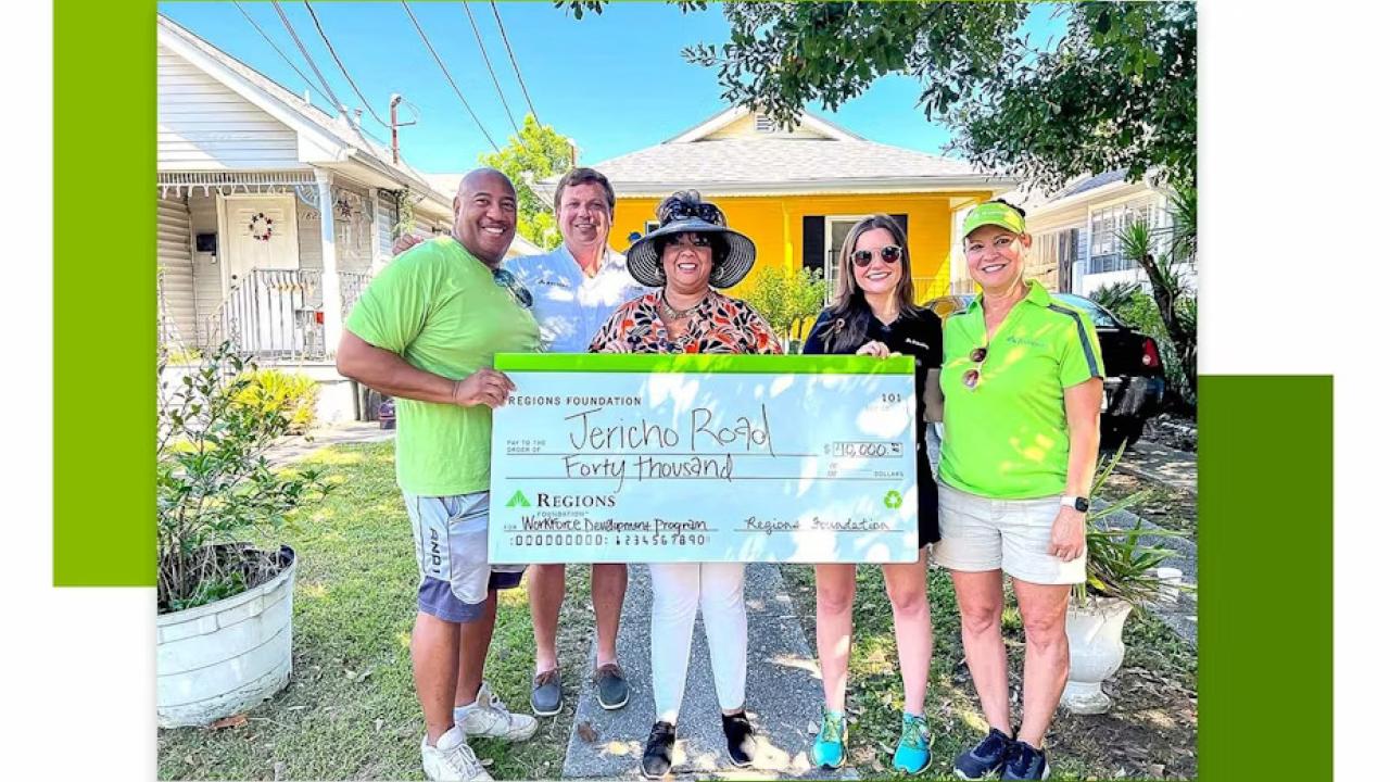 Five people holding a large check in front of a yellow house.