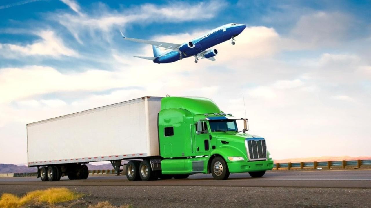 A green semi truck on the road and a jet plane in the sky above it.