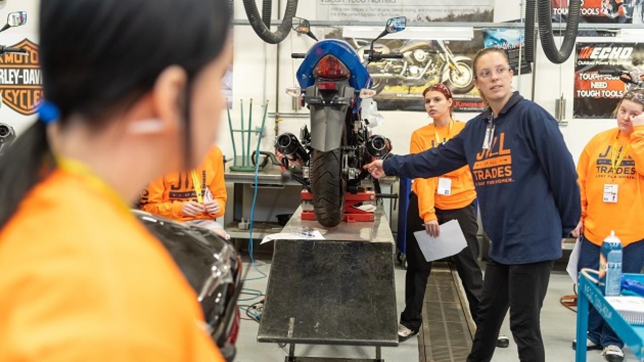 An adult pointing to a spot on a raised motorcycle as students look on.