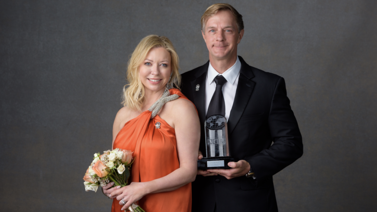 Two people stood next to each other smiling, one person holding flowers and the other holding an award