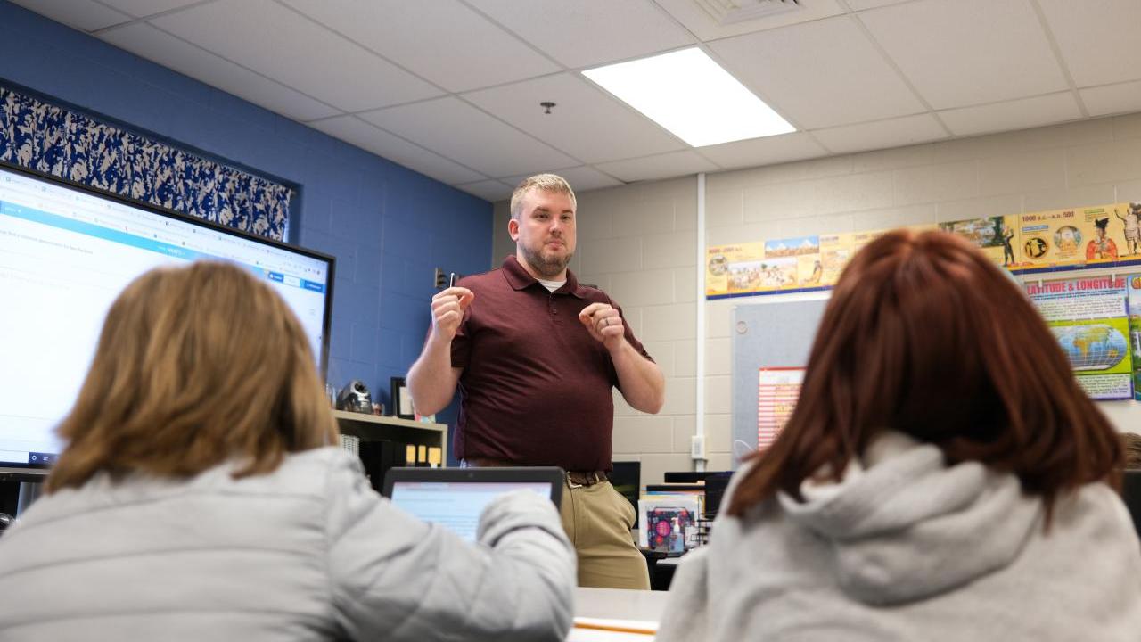 Classroom with students sat at a desk while a teacher is stood in front of the class 