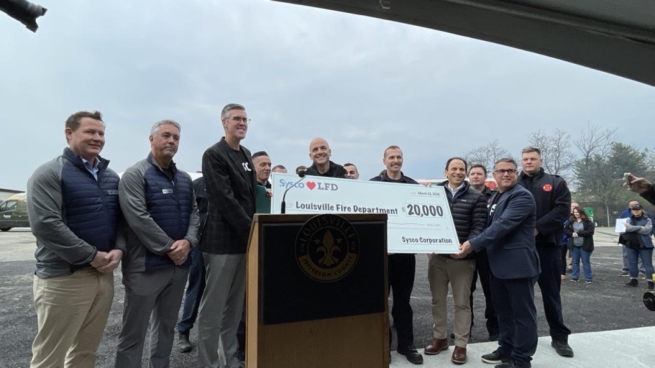 A group of people posed with a large check behind a podium outside.