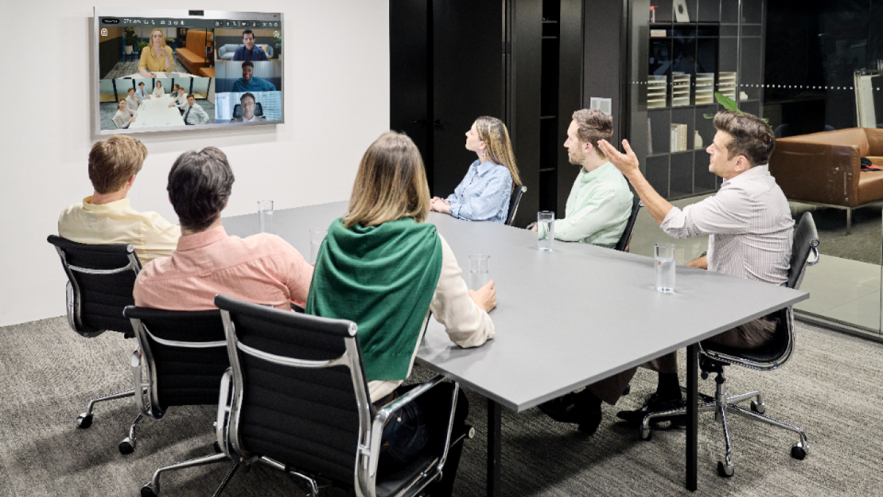 employees seated around a conference table viewing other team members on a monitor mounted on a wall 