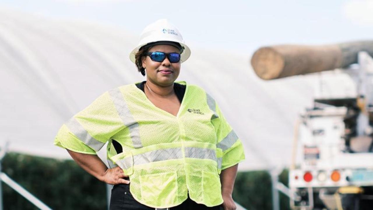 LaQuitta Ghent next to large equipment, wearing high-vis vest at hard hat. One hand on a hip.