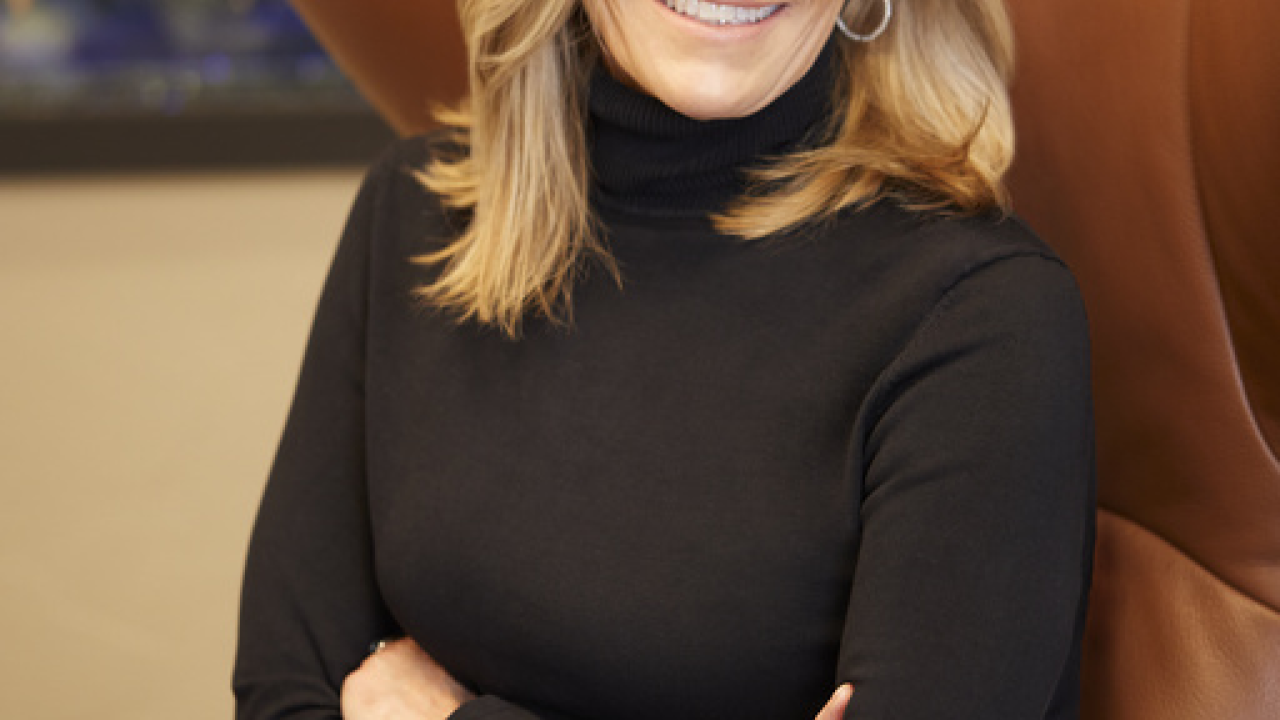 Laura Kohler sitting in a wingback leather chair, smiling, arms crossed, wearing a black turtleneck and pants.