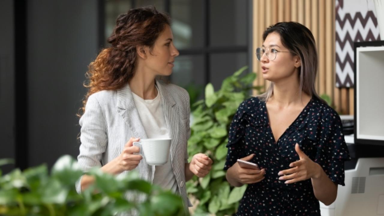 Two people talking, one with a white cup in their hand, the other has a phone. In an office setting with house plants in front and behind.