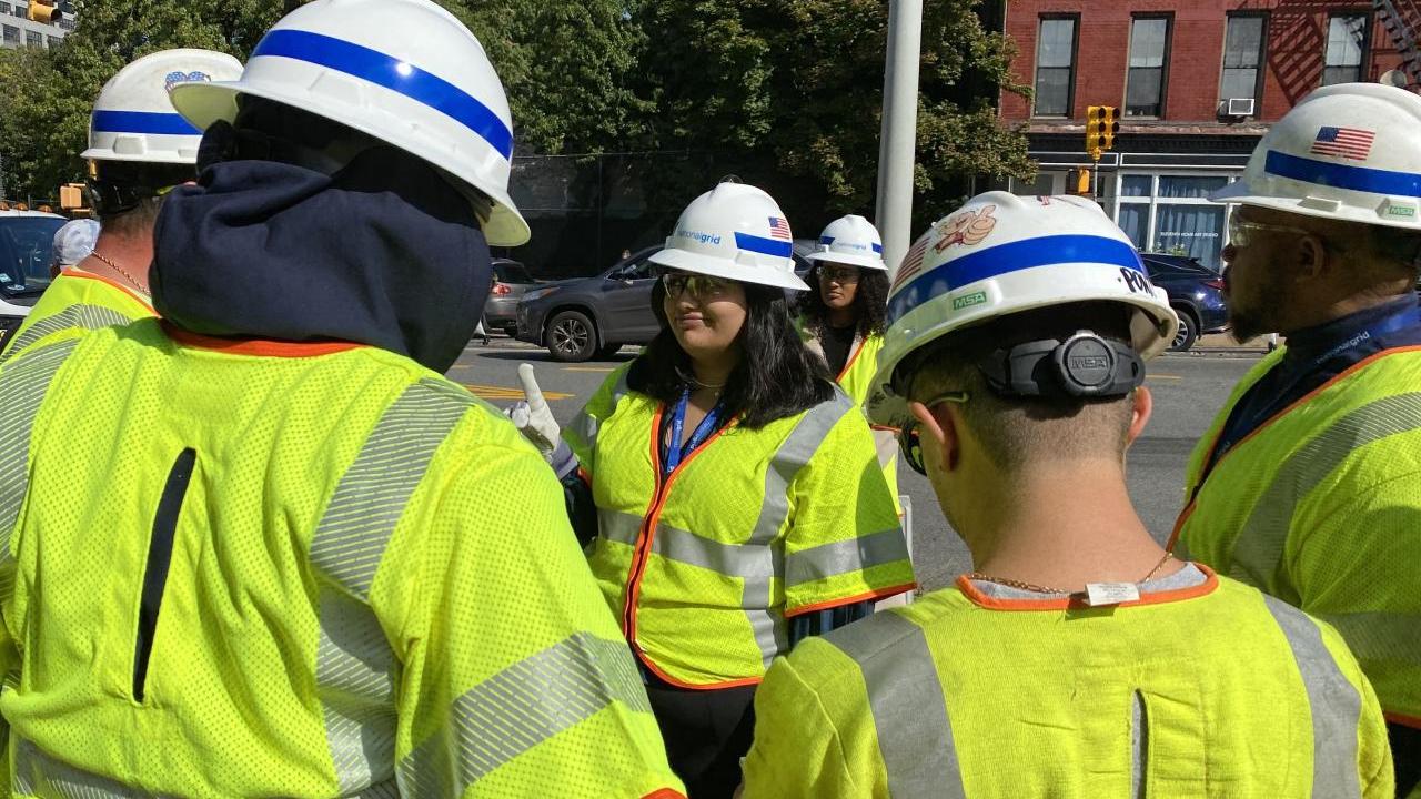 Girl stands on street corner with staff from National Grid in yellow safety vests.