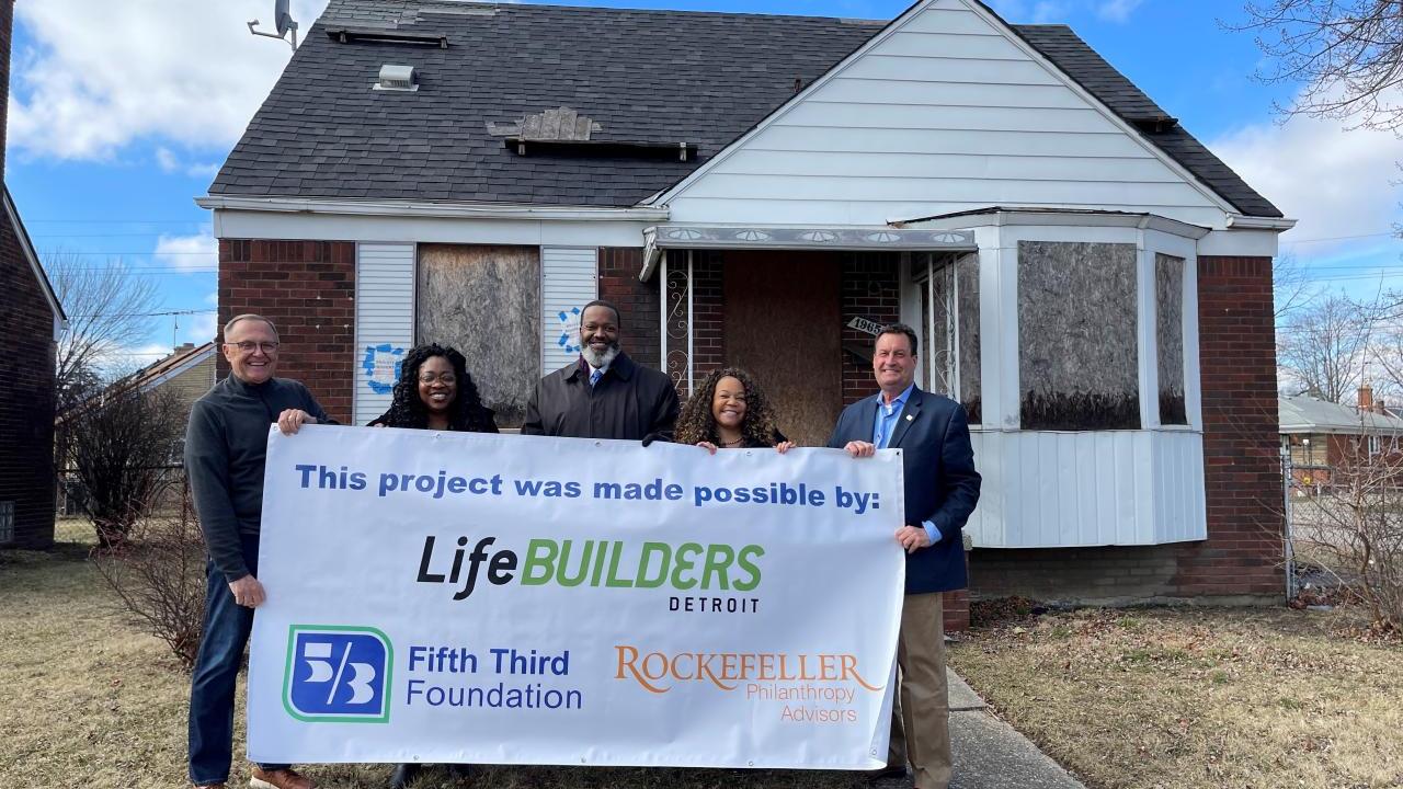 5 people holding a LifeBUILDERS sign outside a house