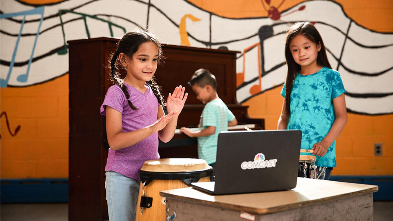 children standing in front of a laptop, one behind them playing on a piano. Wall behind them with a painted mural of floating music notation.