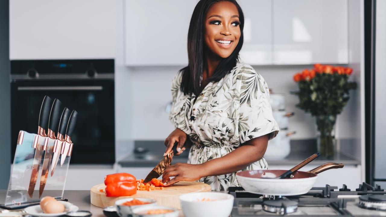 A photo of Celebrity Chef Lorna Maseko standing at a kitchen counter, looking over her left shoulder while cutting a vegetable using her new culinary line for HSN. Lorna has long black hair, medium brown skin and is smiling. She is wearing a cream and green accented short-sleeved dress.