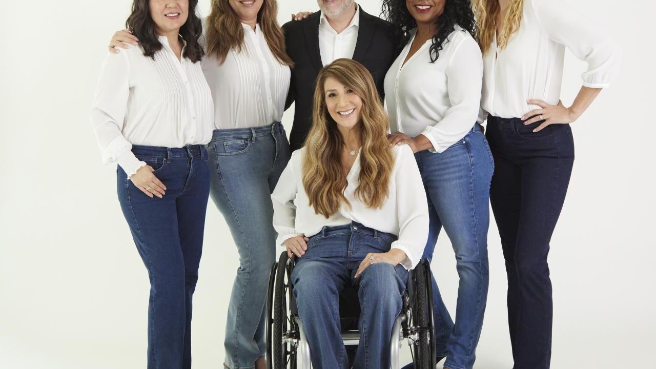 Five diverse models are showcasing NYDJ's Marilyn Straight Adapt-Denim™ jeans alongside Brand Ambassador Mark Peters. All models pose in various denim hues with white tops, while Mark, donning a white shirt and black sportscoat, stands among them. In the forefront, a wheelchair model poses elegantly.