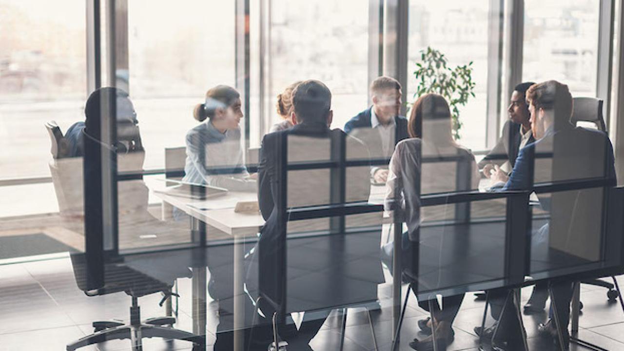 Group of eight men and women of diverse ethnic backgrounds are sitting at a boardroom table.