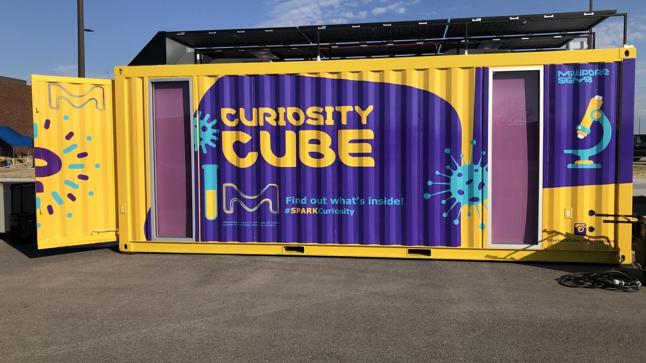 A yellow and purple shipping container with the words "Curiosity Cube" on the side showcase an environment dedicated to expanding science education