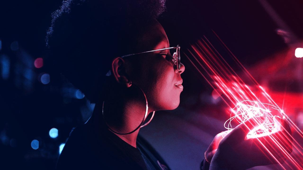Black person with glasses and large earrings smiling with eyes closed, holding glowing pink lights