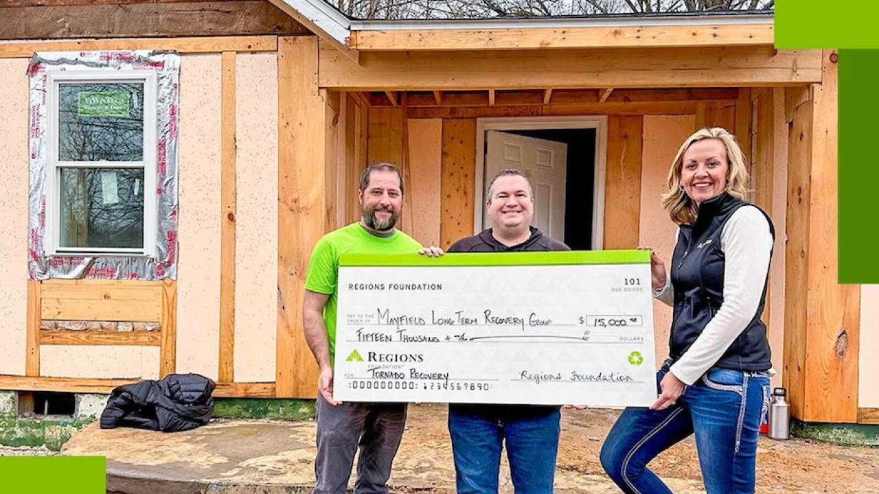 Three people holding a large check, a home being re-built behind them.