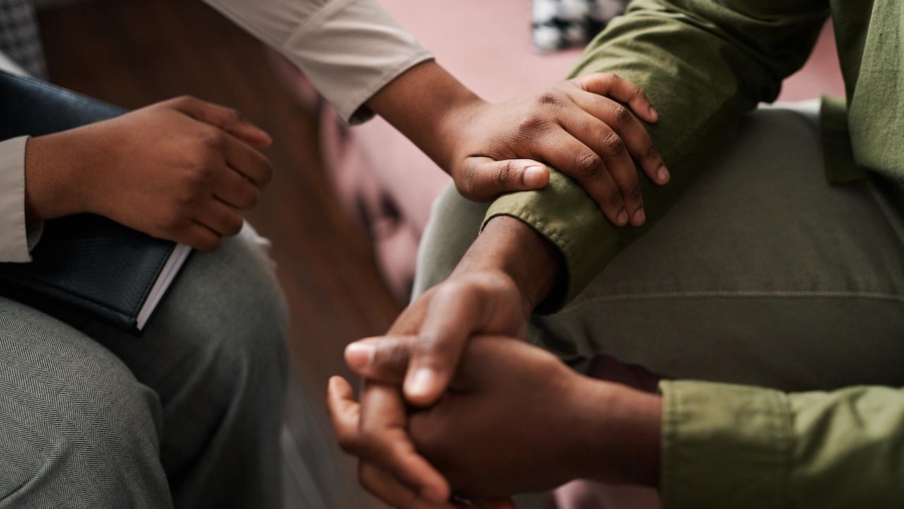 Two people seated. One with folded hands and the other placing his arm in a reassuring manner on the others arm.