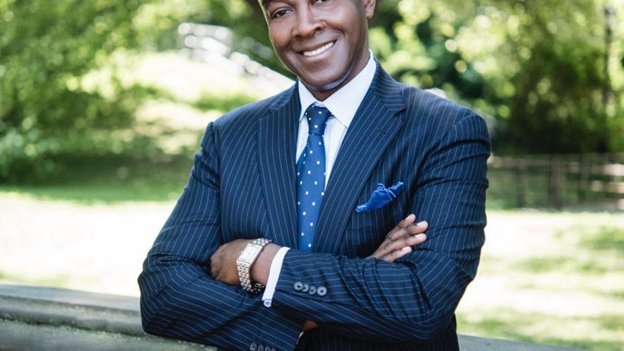 The color photograph shows Michael Carby, of Jamaican heritage leaning against a stone railing located outdoors. He’s wearing a blue pin stripped suit, with a blue polka dot tie, a blue handkerchief and a watch. His arms are crossed and he’s smiling at the camera. It’s a sunny day with green grass and green trees and bushes in the background.