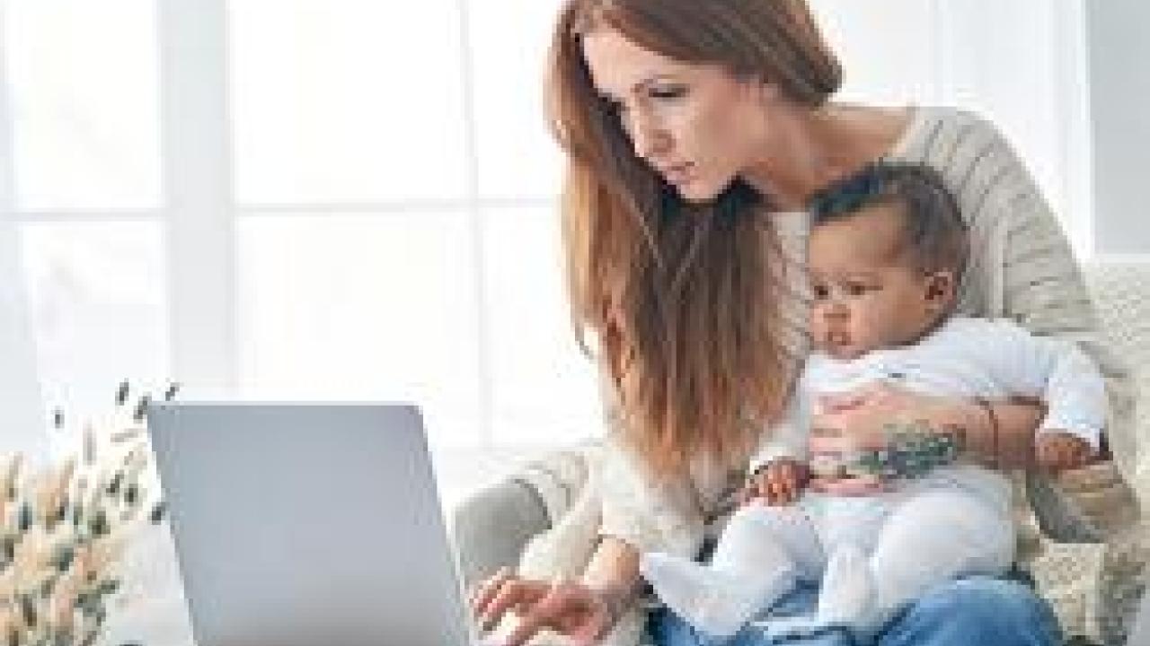 person holds a baby on their lap while working at a laptop in a home setting